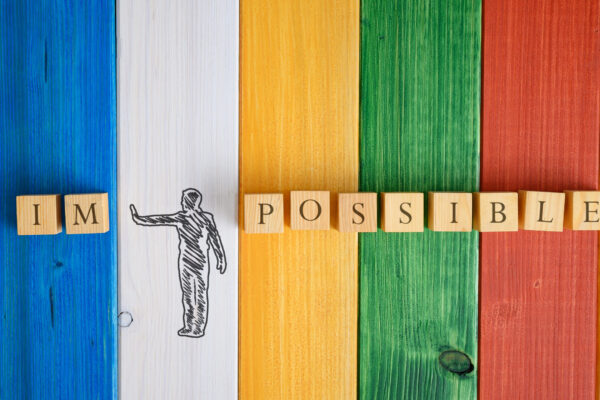 Square wooden pieces arranged on a colourful wood plank background, forming the word 'impossible,' with a depiction of a person pushing the 'im' away.
